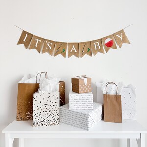 May include: A baby shower scene with a banner that reads "IT'S A BABY". The banner is made of burlap and has fishing-themed decorations. Gift bags and wrapped presents are arranged on a white table, creating a festive atmosphere.