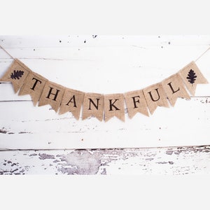 May include: A burlap banner with the word "THANKFUL" in black letters and two brown leaf accents on a whitewashed wood background.