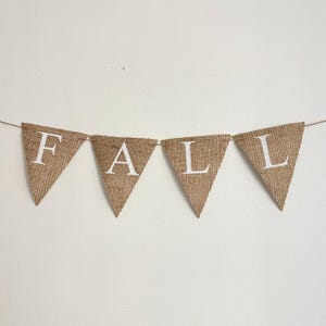 Może przedstawiać: Baner z juty z napisem "FALL" napisanym białymi literami. Baner jest zawieszony na białej ścianie.
