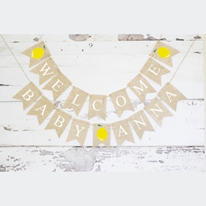 May include: A burlap banner with the words "Welcome Baby Anna" in white letters. The banner is decorated with yellow lemons with green leaves.