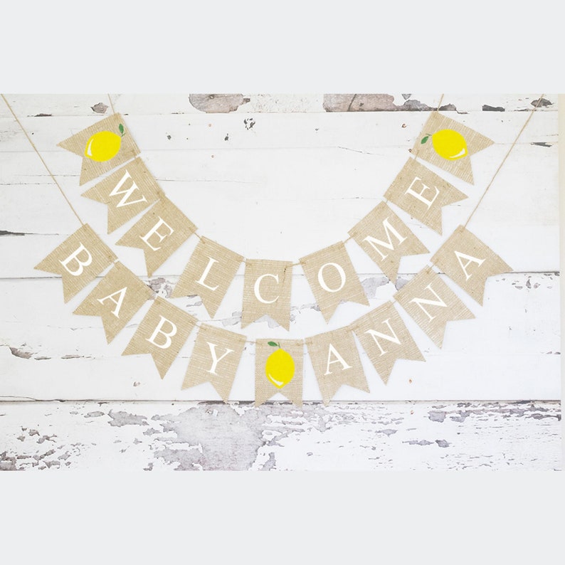 May include: A burlap banner with the words "Welcome Baby Anna" in white letters. The banner is decorated with yellow lemons with green leaves.