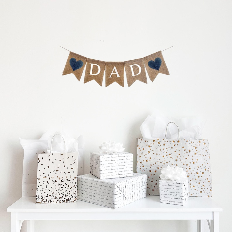 May include: A burlap banner with the word "DAD" in white letters and blue hearts on each end. The banner is hanging on a white wall above a table with several gift bags and boxes wrapped in white paper with gold accents.