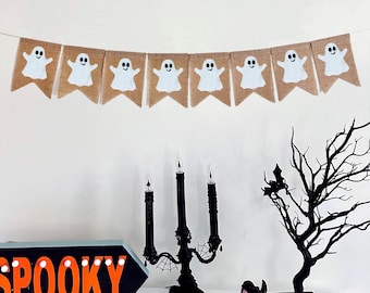 Op de afbeelding: Een Halloween-banner met zeven jute vlaggetjes, elk met een wit spookontwerp. Daaronder een zwarte kandelaar met drie nepkaarsen, een zwarte kraai en een decoratieve zwarte boom. Een bord met het woord "SPOOKY" is ook zichtbaar.