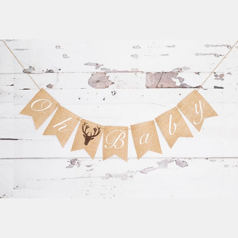 May include: A burlap banner with the words "Oh Baby" in white lettering. A brown deer silhouette is on the "B" flag. The banner is strung on a whitewashed wood background.