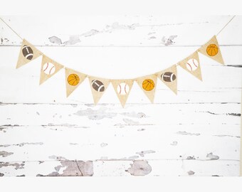 May include: A burlap banner with a string of triangular flags featuring different sports balls, including a baseball, basketball, and football. The flags are strung together with twine and are hanging against a white wooden background.
