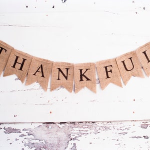 May include: A burlap banner with the word "THANKFUL" printed in black letters. The banner is decorated with two brown leaf shapes on either end.