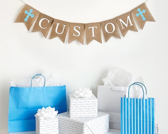 Puede incluir: Una pancarta de arpillera con la palabra "CUSTOM" en letras blancas y cruces azules. Debajo, bolsas de regalo y regalos envueltos en papel azul, blanco y estampado se encuentran sobre una superficie blanca, creando una escena festiva.