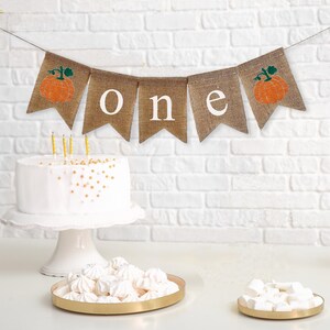 May include: A burlap banner with the word "one" in white letters and two orange pumpkins with green stems. The banner is strung on a white wall above a white cake with gold sprinkles and a white cake stand. There are two gold trays with white meringue cookies on a white surface.