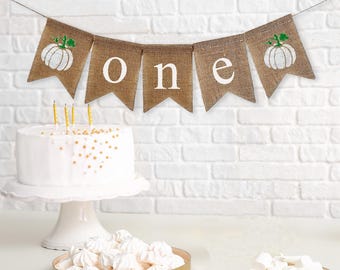May include: A burlap banner with white pumpkins and the word "one" in white letters. The banner is strung up on a white brick wall. There is a white cake with gold sprinkles on a white cake stand in the foreground.