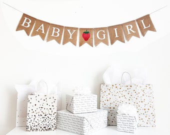 May include: A burlap banner with the words "BABY GIRL" in white letters and a red strawberry graphic. The banner is hanging from a white wall above a white table with four gift bags. The gift bags are wrapped in white paper with different patterns, including gold stars, gold confetti, and a repeating pattern of the words "Love you more" in gold lettering.