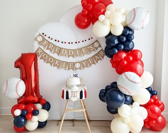 Pode incluir: Decoração de festa de primeiro aniversário com tema de beisebol. Apresenta um balão vermelho com o número um, balões em forma de bola de beisebol e uma faixa que diz "Freddie's Rookie Year". Um pequeno bolo está em uma cadeira alta decorada com tema de beisebol.