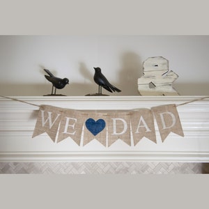 Pu&ograve; includere: Uno striscione di juta con le parole "WE &hearts; DAD" in lettere bianche. Il cuore &egrave; blu. Lo striscione &egrave; appeso a un camino bianco con due statuine di uccelli neri e un simbolo di ampersand in legno bianco.