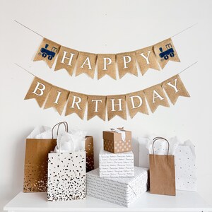 May include: A burlap banner with the words "Happy Birthday" and two blue train images. The banner is hanging on a white wall above three brown paper gift bags and a white gift bag with black confetti. The gift bags are on a white table.