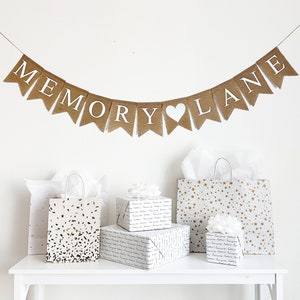 May include: A banner that reads "MEMORY LANE" hangs above gifts and gift bags. The banner is made of burlap with white letters and a white heart. The gifts are wrapped in patterned paper, and the gift bags have various designs.