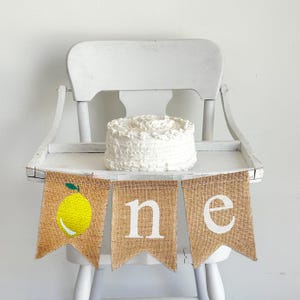 A burlap banner made from three 5 by 7 inch fishtail shaped flags. The first flag has a lemon image on it. The second flag has a lowercase N in white, and the third flag has a lowercase E in white. It is shown on a high chair in the photo.