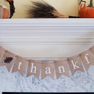 May include: A burlap banner with the words "be thankful" in white letters. The banner is hanging from a white mantel above a marble fireplace.