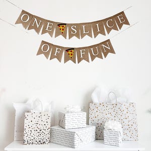 May include: A festive scene with a banner reading "ONE SLICE OF FUN" above gift bags and wrapped presents. The banner is brown with white letters and pizza slice graphics. Gift bags and presents are decorated with confetti, stars, and handwritten text.