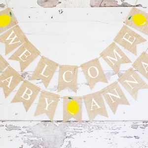 May include: A burlap banner with the words "Welcome Baby Anna" in white letters. Each letter is on a separate flag with a small yellow lemon with a green leaf on top. The banner is strung on a white background.