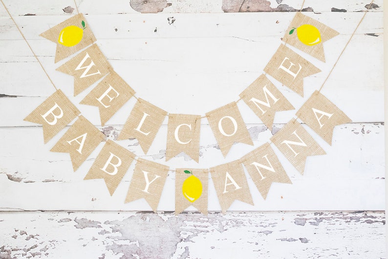 May include: A burlap banner with the words "Welcome Baby Anna" in white letters. Each letter is on a separate flag with a small yellow lemon with a green leaf on top. The banner is strung on a white background.
