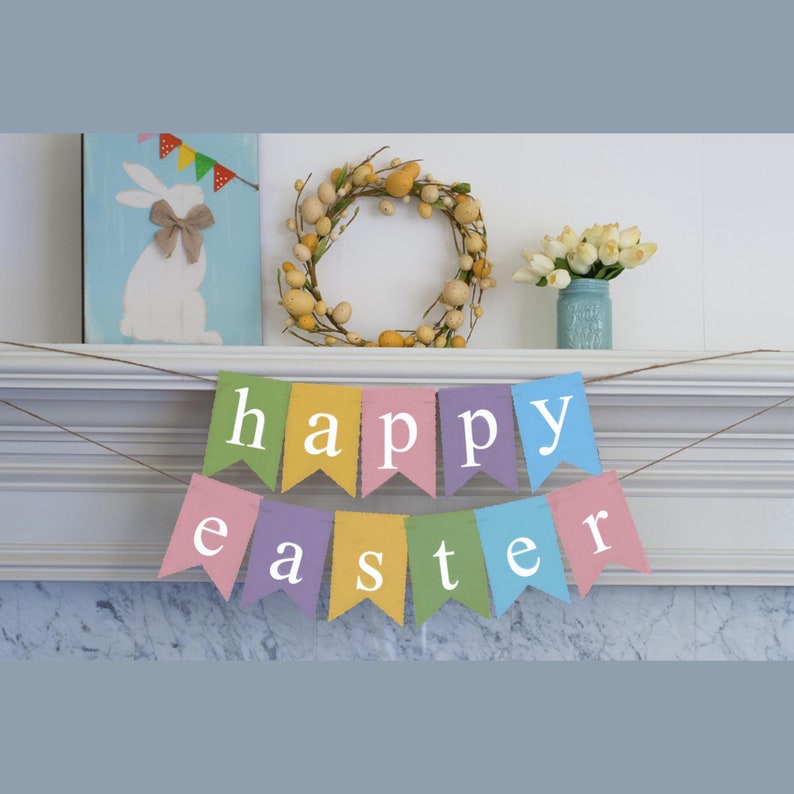 May include: A colorful banner with the words "happy easter" in white letters on a light blue background. The banner is strung on a white mantel with a wreath, a painting of a bunny, and a vase of white flowers.