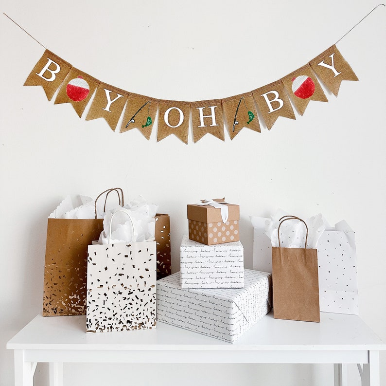 May include: A banner that reads "BOY OH BOY" with fishing bobber and fishing rod illustrations hangs above gift bags and wrapped presents. The gifts are on a white table, with a white wall in the background.