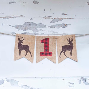 May include: A burlap banner with a red and black plaid number "1" in the center, flanked by two brown deer silhouettes. The banner is strung with twine and hangs against a white, weathered wood background.