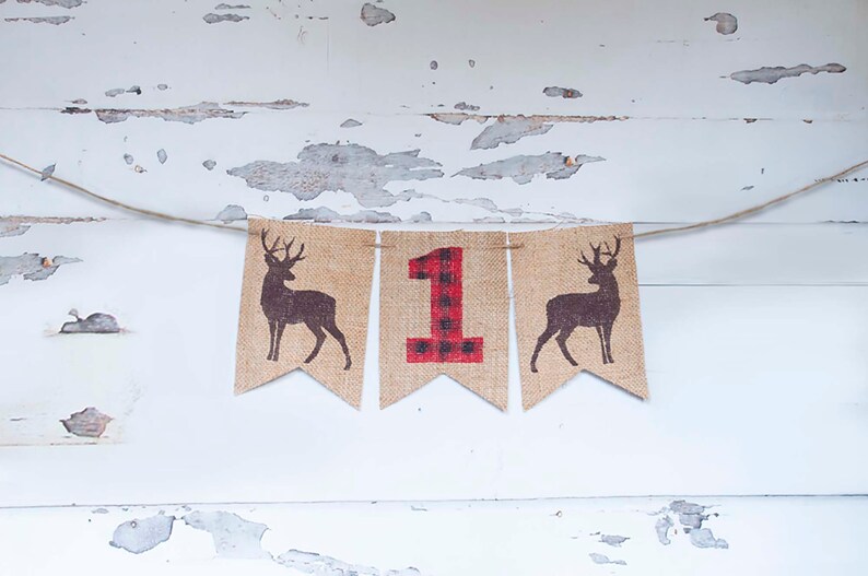 May include: A burlap banner with a red and black plaid number "1" in the center, flanked by two brown deer silhouettes. The banner is strung with twine and hangs against a white, weathered wood background.