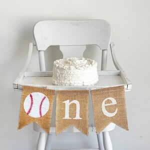 May include: A burlap banner with the word "one" and a baseball graphic. The banner is hanging on a white high chair with a white cake on the tray.