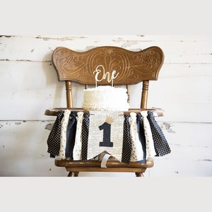 May include: A white cake with a gold 'one' topper sits on a high chair with a black, white, and gold fabric banner that says '1' with a gold crown.