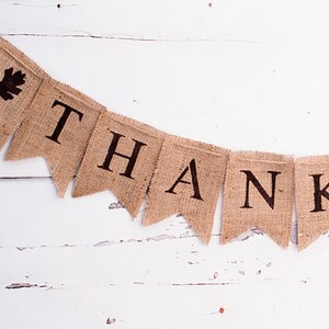 May include: A burlap banner with the word "THANK" spelled out in black letters. The banner is strung on a white wooden surface.