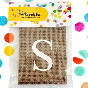 May include: A burlap banner with the letter "S" printed in white. The banner is packaged in a clear plastic bag with a colorful confetti design.
