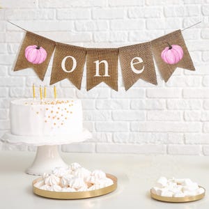 May include: A burlap banner with pink pumpkins and the word "one" in white letters. The banner is hanging above a white cake with gold sprinkles and three lit candles.