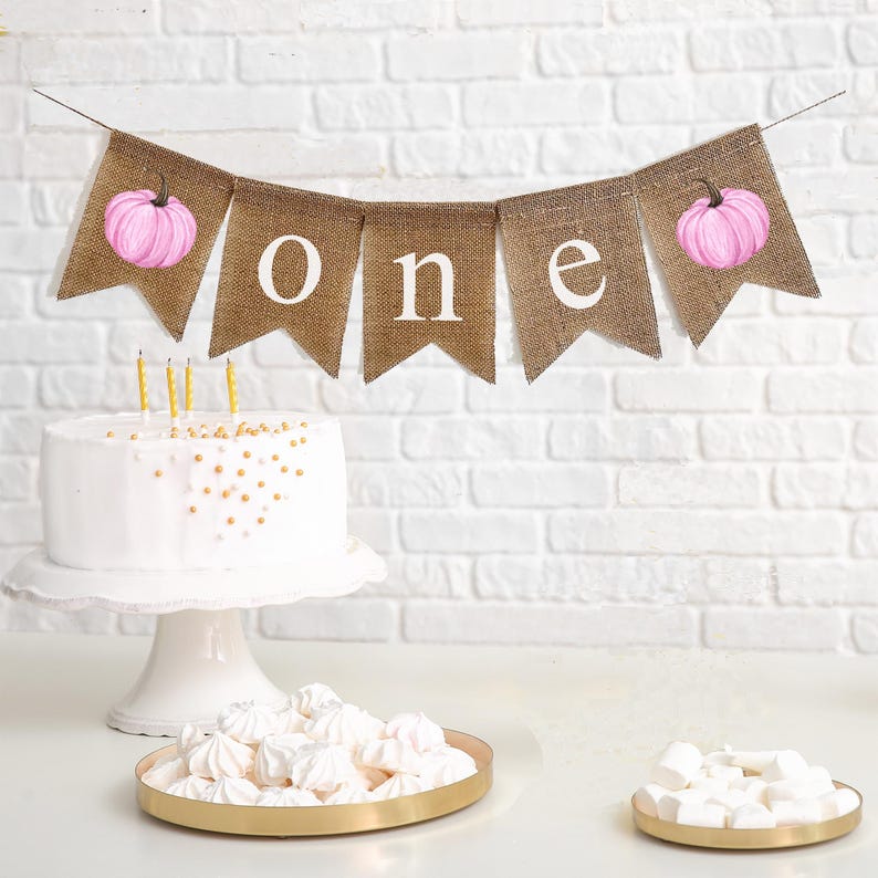 May include: A burlap banner with pink pumpkins and the word "one" in white letters. The banner is hanging above a white cake with gold sprinkles and three lit candles.