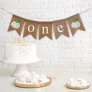 May include: A burlap banner with white pumpkins and the word "one" in white letters. The banner is strung up on a white brick wall. There is a white cake with gold sprinkles on a white cake stand in the foreground.