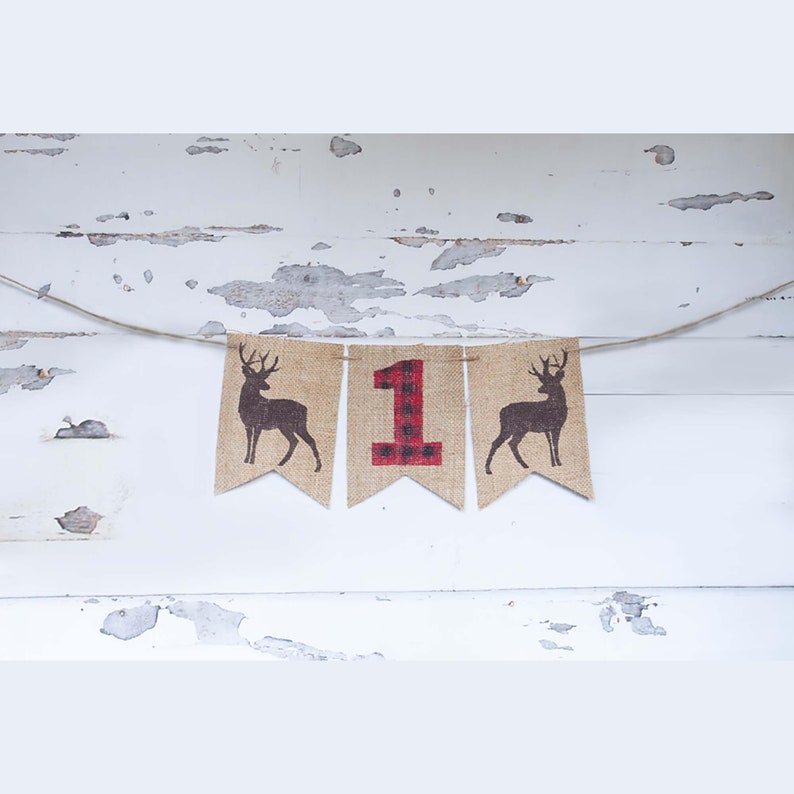 May include: A burlap banner with a red and black plaid number "1" in the center, flanked by two brown silhouette deer on each side. The banner is strung with twine.