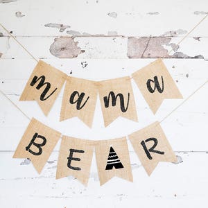 May include: A burlap banner with the words "mama bear" in black lettering. The banner is strung on a white string and hung on a white wooden wall.