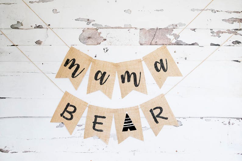 May include: A burlap banner with the words "mama bear" in black lettering. The banner is strung on a white string and hung on a white wooden wall.