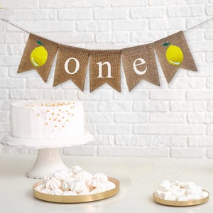 Puede incluir: Una pancarta de arpillera con la palabra "one" en letras blancas e ilustraciones de limones. La pancarta est&aacute; colgada sobre un pastel blanco y una pared de ladrillo blanco.