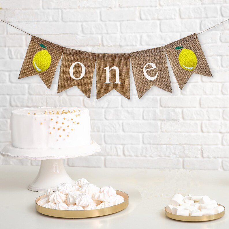 Puede incluir: Una pancarta de arpillera con la palabra "one" en letras blancas e ilustraciones de limones. La pancarta est&aacute; colgada sobre un pastel blanco y una pared de ladrillo blanco.