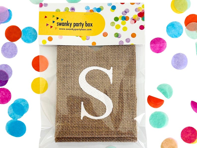 Op de afbeelding: Een jute zak met een grote witte letter "S" op de voorkant. De zak is verpakt in doorzichtig plastic. De bovenkant van de verpakking heeft een gele banner met de tekst "swanky party box" en kleurrijke confetti.