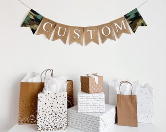 May include: A banner that says "CUSTOM" hangs above gift bags and wrapped presents. The banner has a camouflage pattern on the ends. The gifts are on a white table. The gift bags have various patterns.