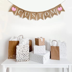 Puede incluir: Una exhibición festiva con una pancarta de arpillera con la palabra "CUSTOM" y acentos de calabaza rosa. Bolsas de regalo y regalos envueltos en varios tamaños y patrones están dispuestos sobre una mesa blanca.