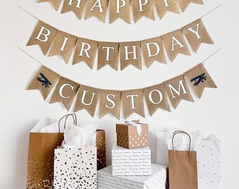 Może przedstawiać: Baner z płótna jutowego z napisem "Happy Birthday Custom" i niebieskimi ilustracjami samolotów. Baner wisi na białej ścianie, pod nim znajdują się brązowe torby prezentowe i papier do pakowania.
