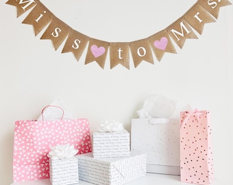 May include: A burlap banner with the text "Miss to Mrs." in white letters and pink glitter hearts. The banner is hanging on a white wall. There are several gift bags and boxes on a white table below the banner.