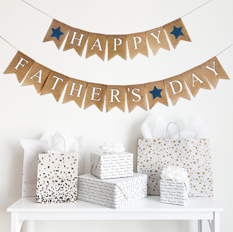 A burlap banner made from 4.5 by 6.5 inch fishtail shaped flags. Each flag has a pocket across the top for stringing. The top line of this banner says HAPPY in white capital letters. There is an image of a navy star on each end of the word. The bottom line says FATHER'S DAY. There is an image of a navy star between the words. It is strung on two 110 inch jute strings.