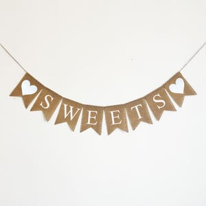 May include: A burlap banner with the word "SWEETS" in white letters and two white hearts on either end. The banner is strung on a white background.