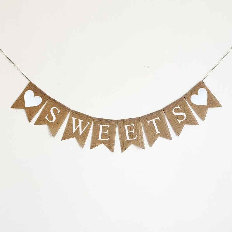 May include: A burlap banner with the word "SWEETS" in white letters and two white hearts on either end. The banner is strung on a white background.
