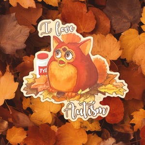 May include: A sticker featuring a cartoon Furby with the text "I love Autism." The Furby is orange and red, with large eyes and a small bottle of medicine. The sticker is set against a background of autumn leaves in shades of orange and brown.