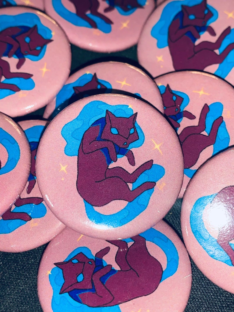 May include: A set of pink and blue button pins featuring a cartoon purple cat curled up in a blue cloud. The cat has a white face and a white tail.