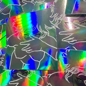 May include: Holographic stickers featuring a stylized illustration of a person with hands covering their face. The artwork is outlined in white against a background that reflects a rainbow of colors. The stickers have a square shape.
